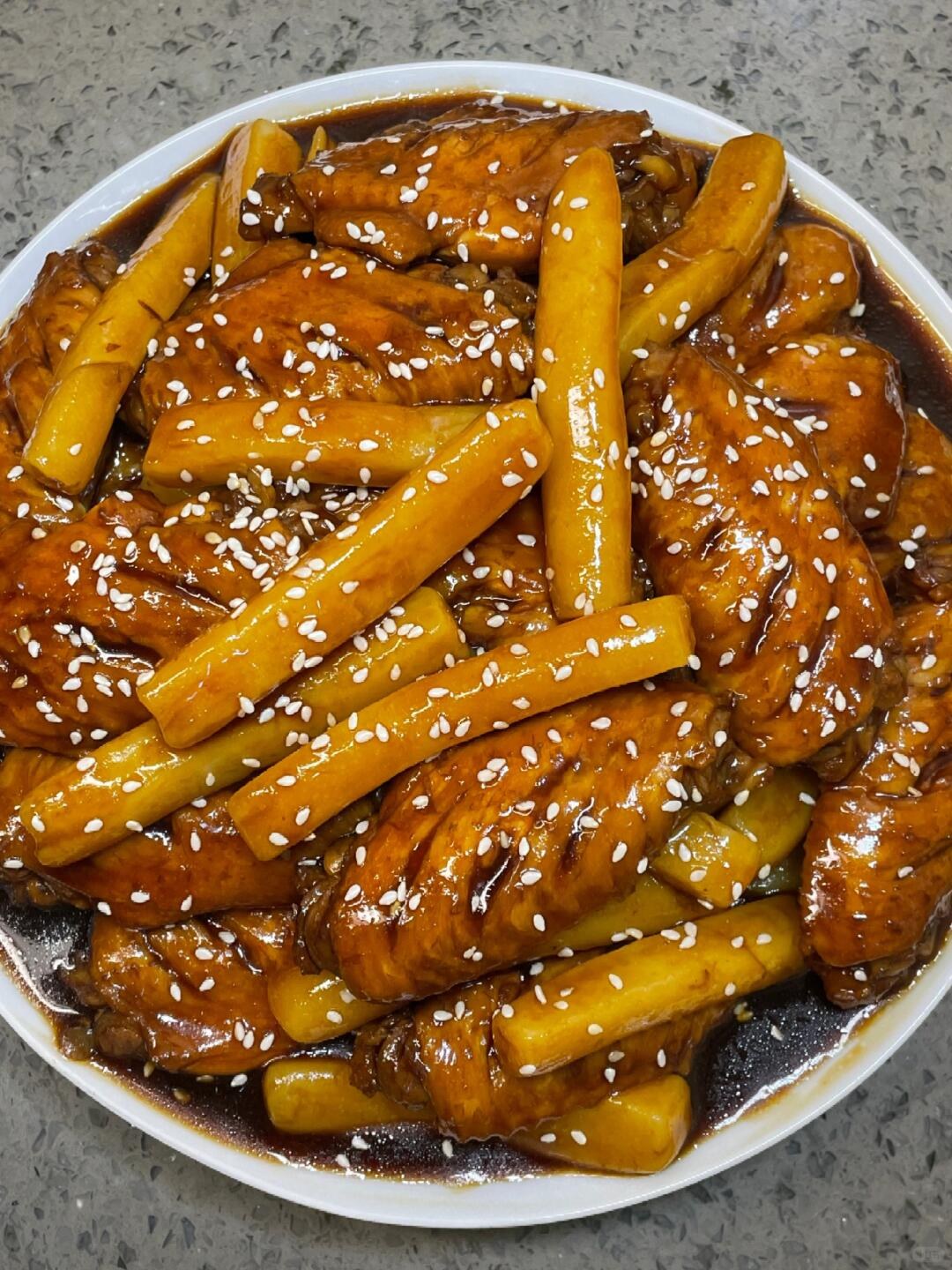 Glossy brown chicken wings and long rice cakes sprinkled with sesame on a white plate.