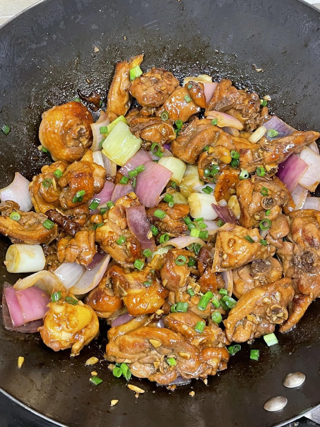 Brown chicken pieces with chopped scallions and purple onion in a dark pan.