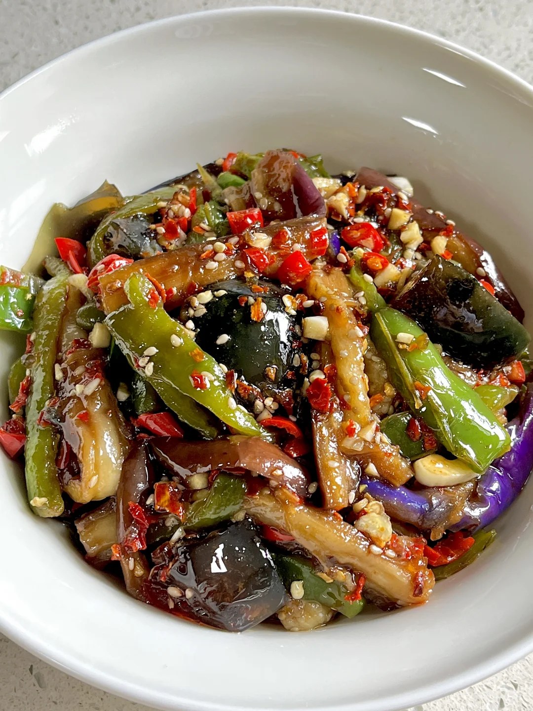 Eggplant strips, dark century egg pieces and green peppers topped with chopped red chiles and sesame in a white bowl.