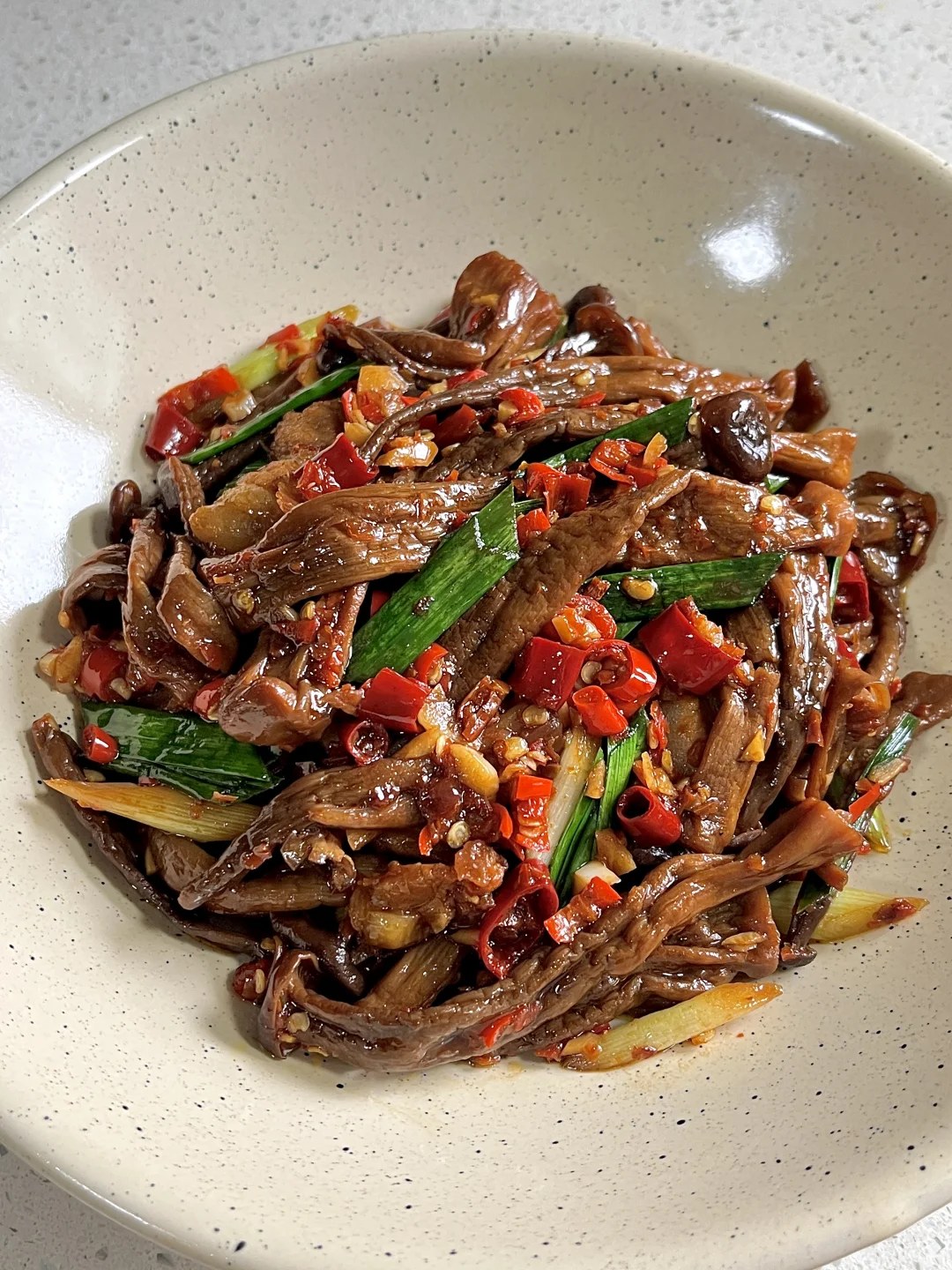 Brown mushrooms and pork pieces with red chiles and green garlic sprouts on a pale plate.