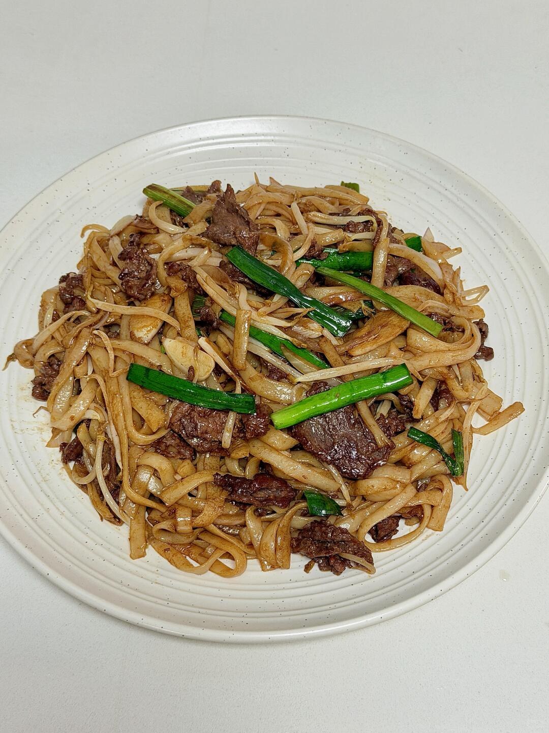 Wide brown rice noodles with beef and green chives on a white plate.