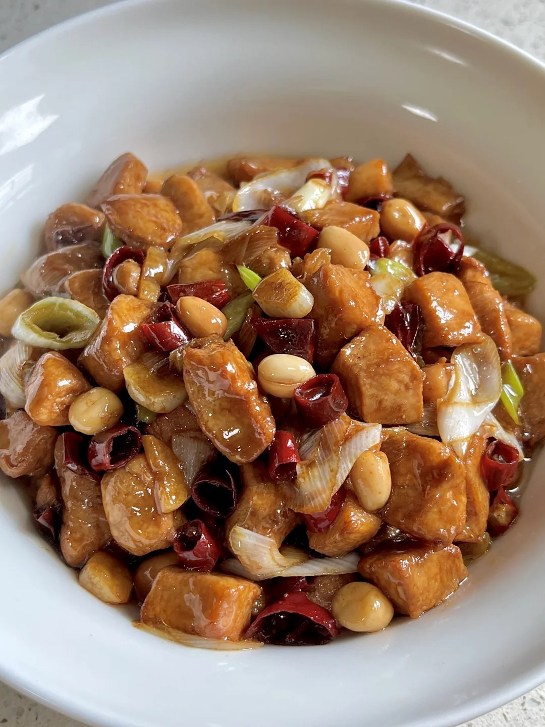 Glazed diced chicken with peanuts, dried red chiles and scallion sections in a white bowl.